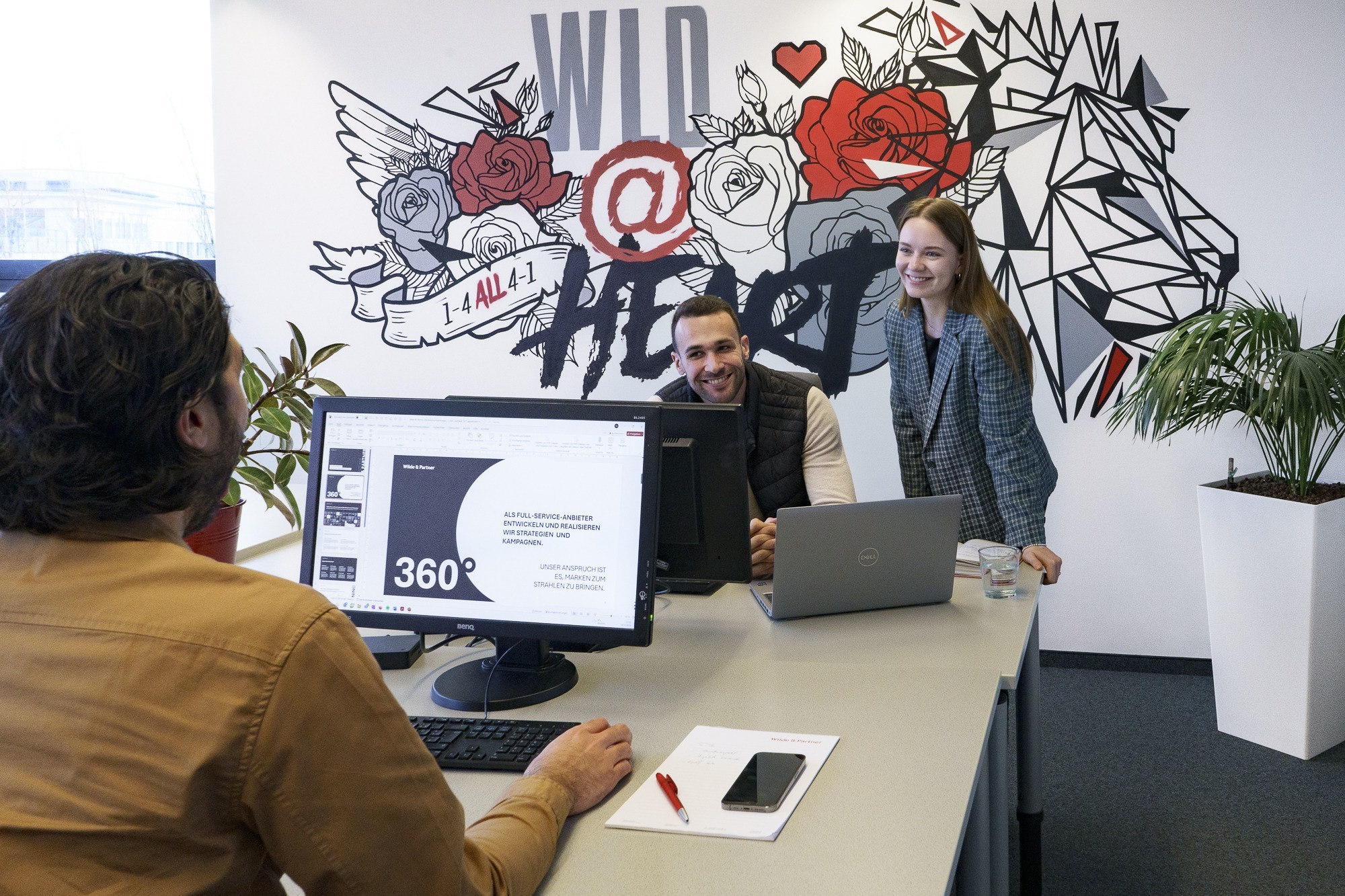 Three people chatting in the office while one is working on a PowerPoint presentation
